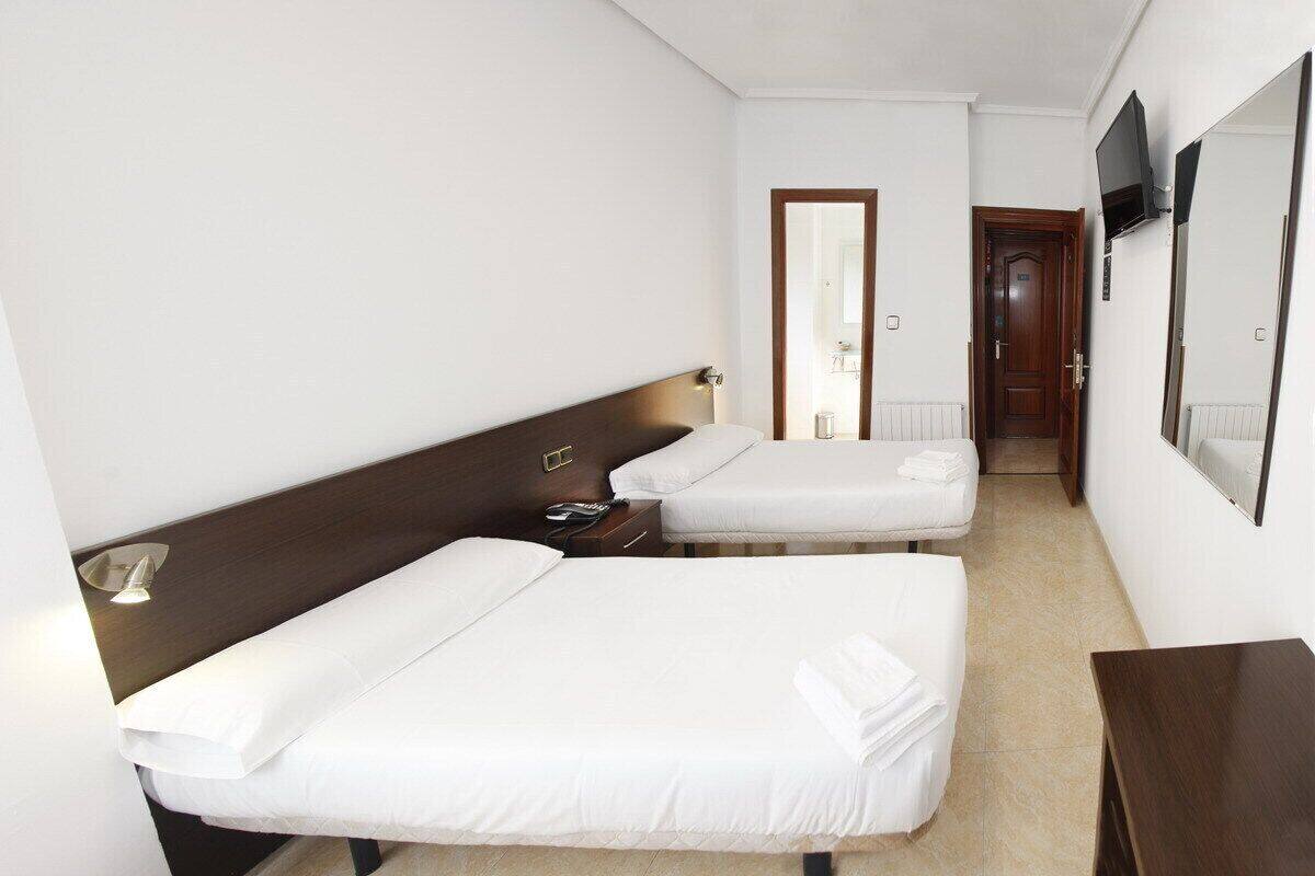 Quarto de hotel com duas camas de solteiro, cabeceira de madeira, mesa de cabeceira com abajur, espelho e televisão na parede.