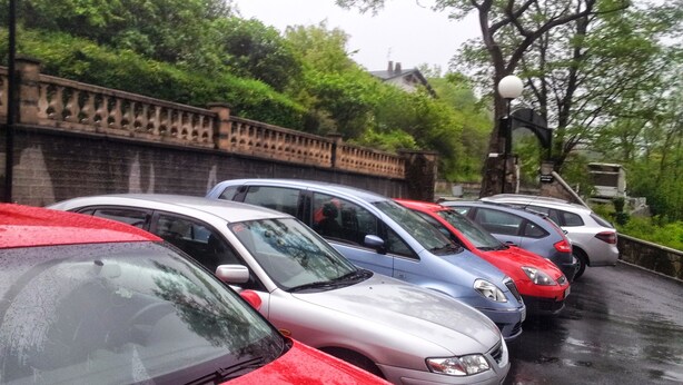 Fila de coches estacionados a lo largo de una carretera mojada, con un muro de ladrillo y vegetación al fondo.