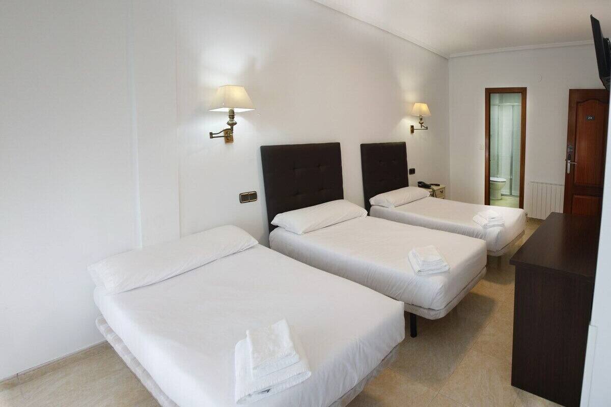 Quarto de hotel com três camas individuais, cada uma com um edredom branco e uma toalha. Há duas luminárias fixadas na parede, uma de cada lado das camas.