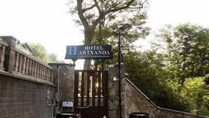 Entrada de un hotel con un letrero que dice "HOTEL ARTXANDA", una pared de piedra, una reja metálica y un gran árbol al fondo.