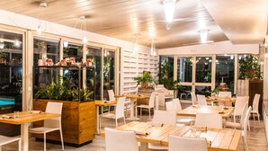 Un restaurante moderno con mesas de madera y sillas blancas, ventanas amplias y plantas de interior.