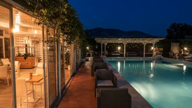 Un área junto a la piscina con sillas de descanso, una piscina y un edificio con puertas de vidrio e iluminación interior.