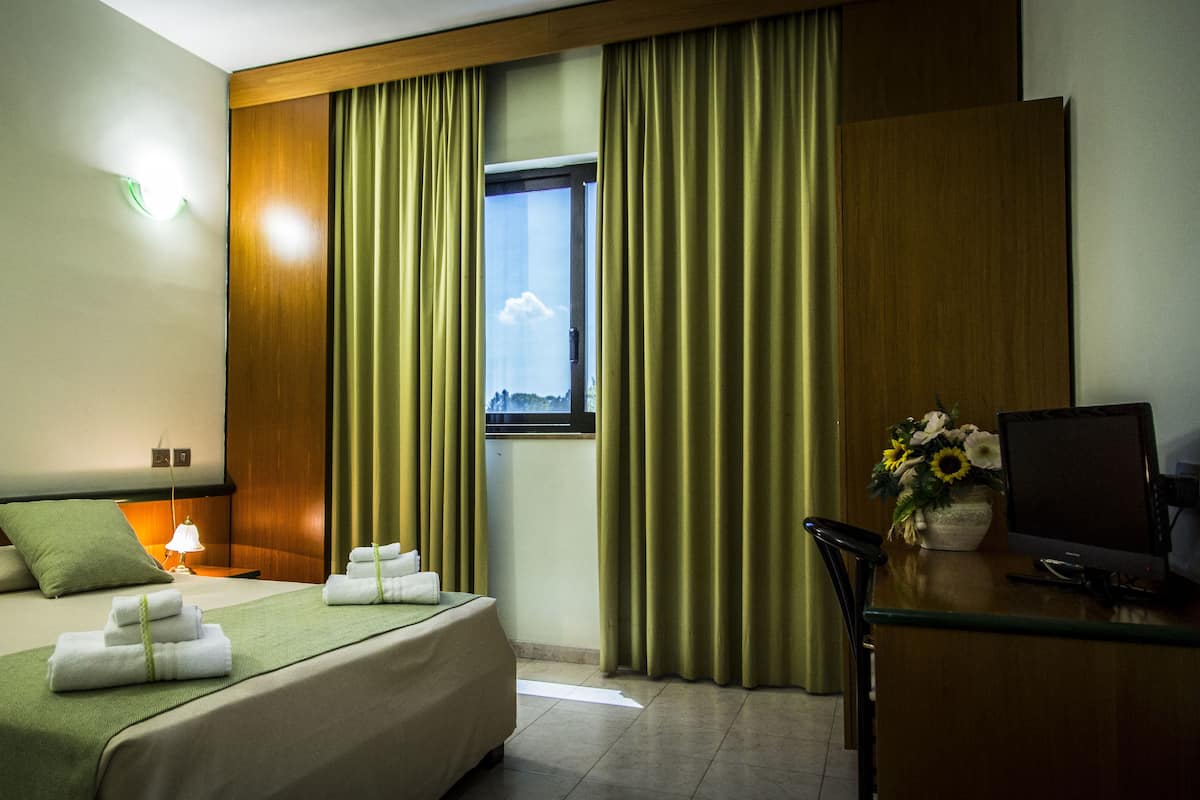 A hotel room with a bed, a desk, a lamp, a window with curtains, and a view of the sky.