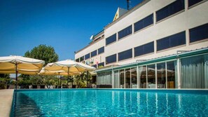 Un hotel con piscina, sombrillas exteriores y un edificio grande con muchas ventanas.