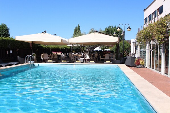 Una piscina con sillas de descanso y sombrillas, rodeada de un patio de ladrillo y un edificio con amplias ventanas.
