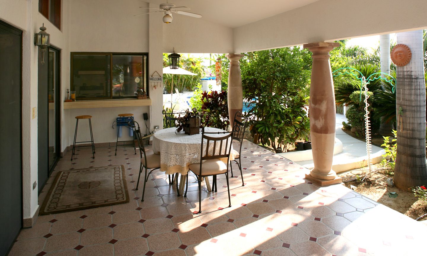 A covered patio with a round table and chairs, a ceiling fan, and a view of a garden with a pool.