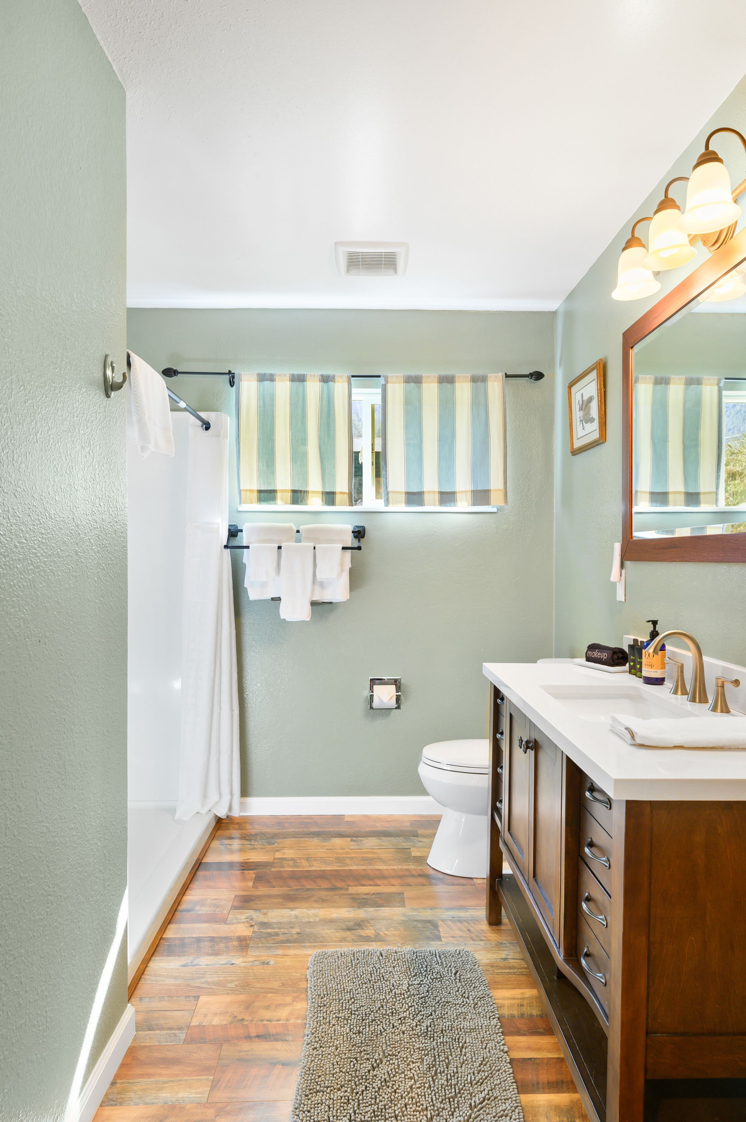 Un bagno con pavimento in legno, un WC bianco, un lavandino bianco con specchio e tende veneziane a righe.