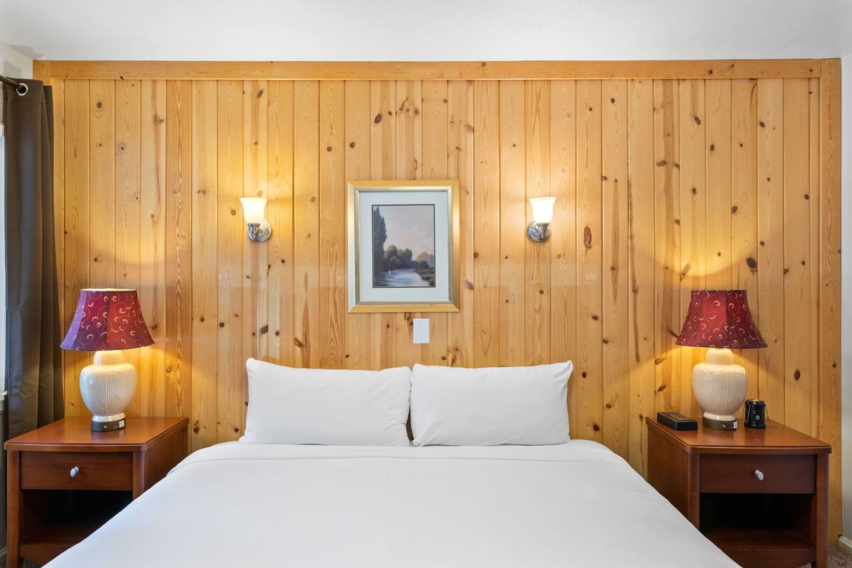Une chambre d’hôtel avec un mur lambrissé en bois, un tableau encadré, deux tables de chevet avec des lampes et un lit avec une literie blanche.