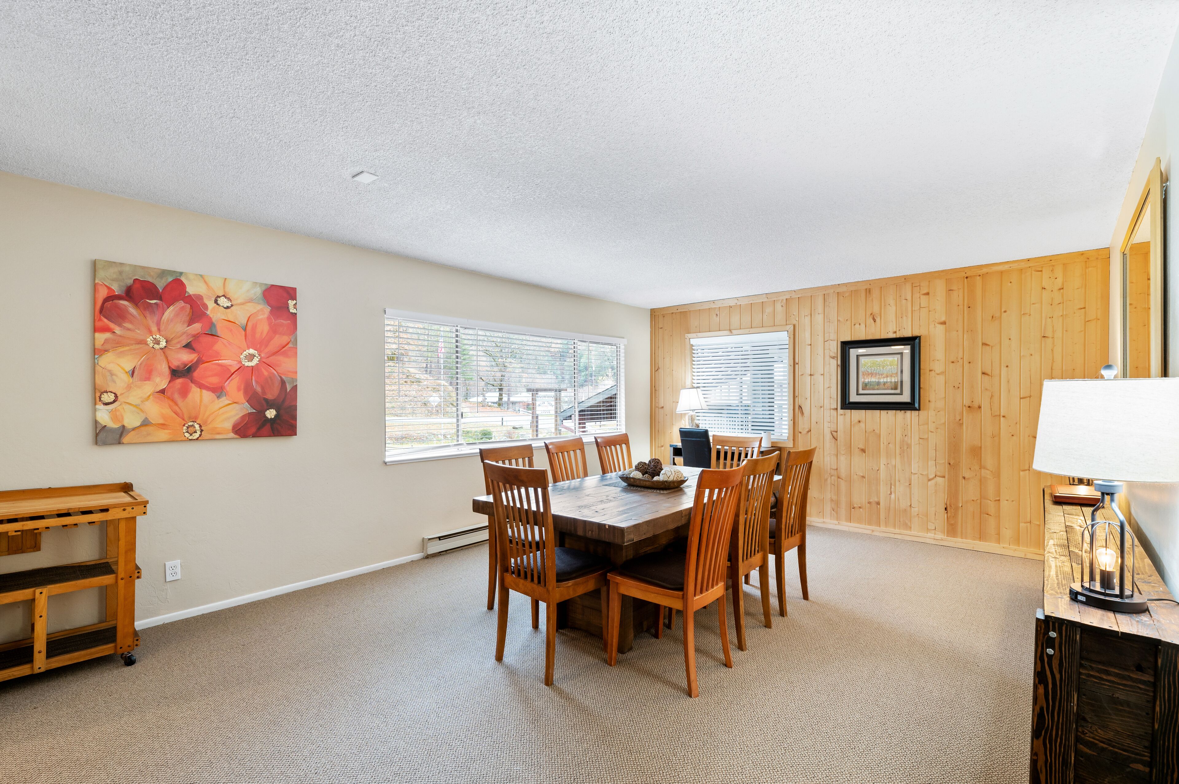A dining area with a wooden table and chairs, a painting on the wall, and a lamp on a side table.