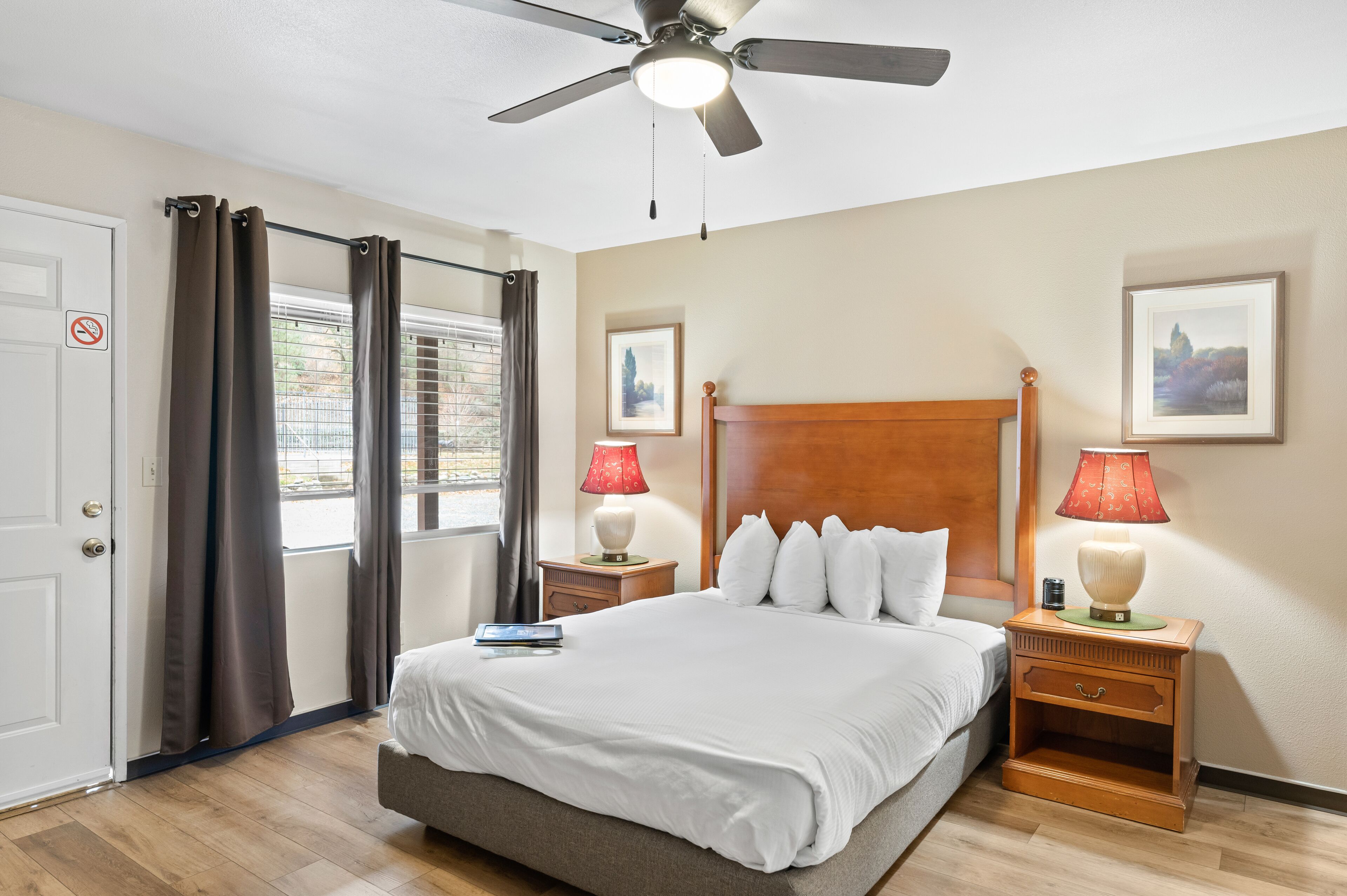A hotel room with a bed, two nightstands, a ceiling fan, and two windows with curtains.