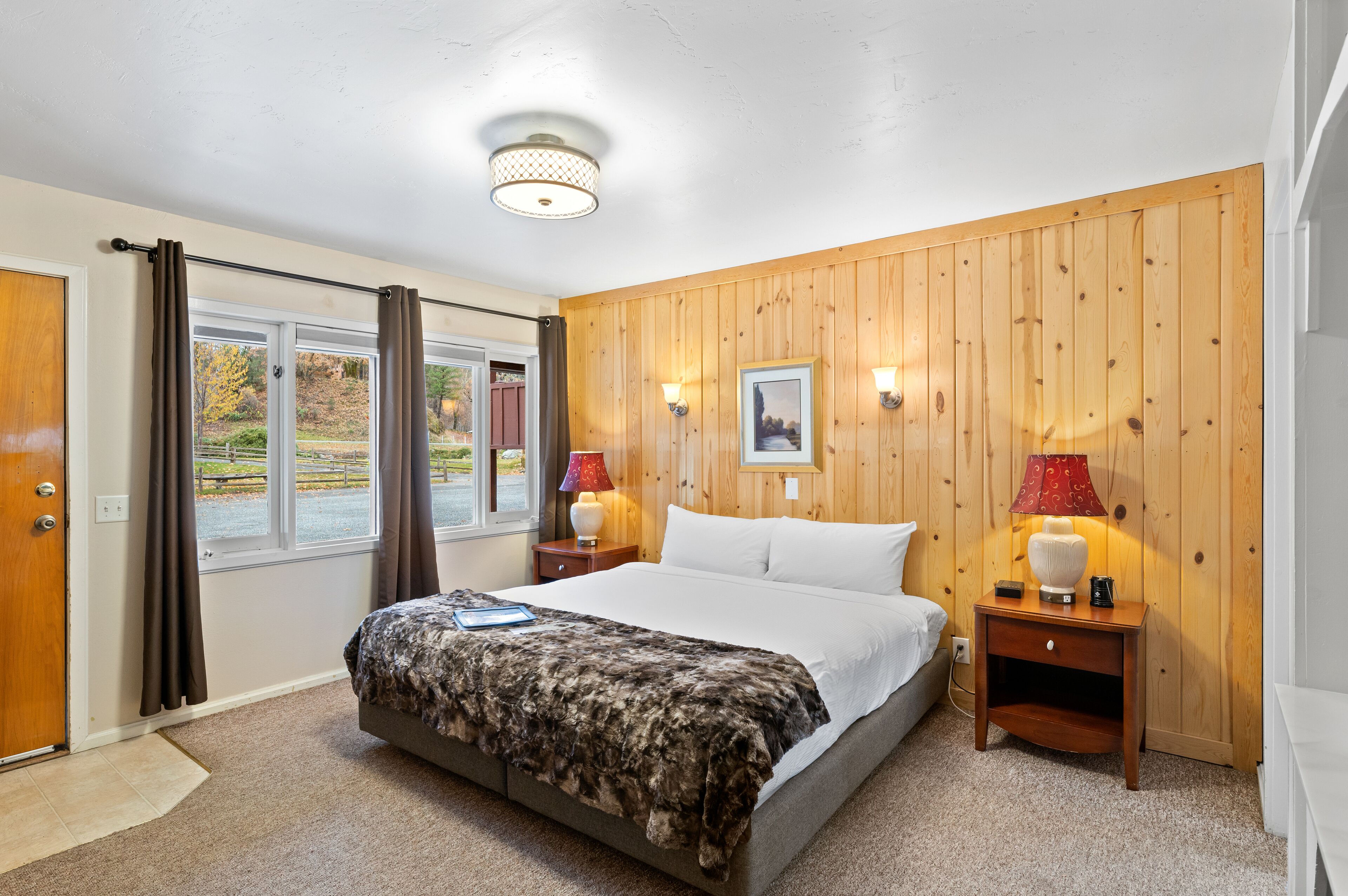 Une chambre à coucher avec un lit, deux tables de chevet avec des lampes, une fenêtre avec des rideaux et un panneau mural en bois.