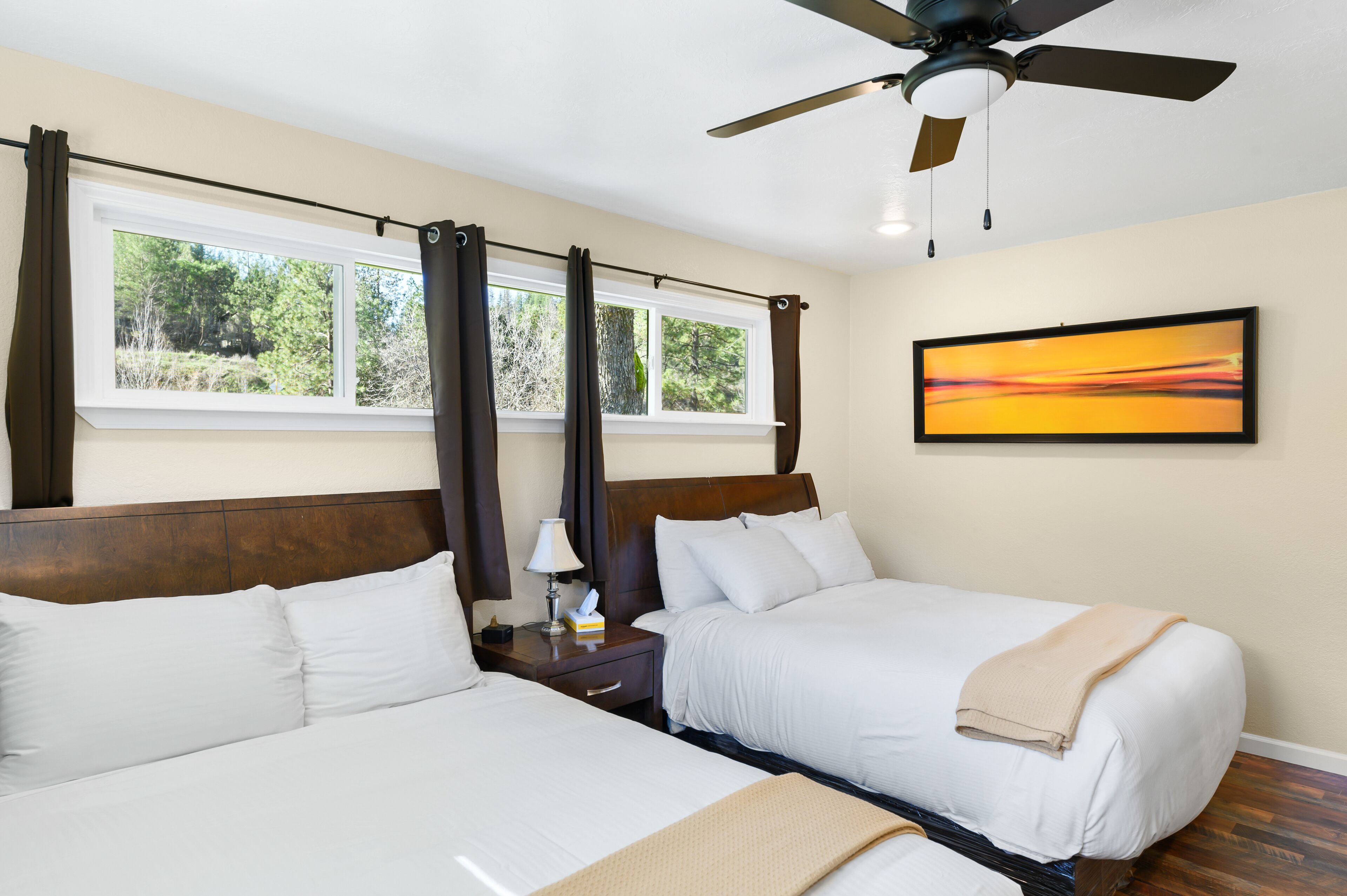 A hotel room with two beds, a ceiling fan, a window with curtains, a bedside table with a lamp, and a painting on the wall.
