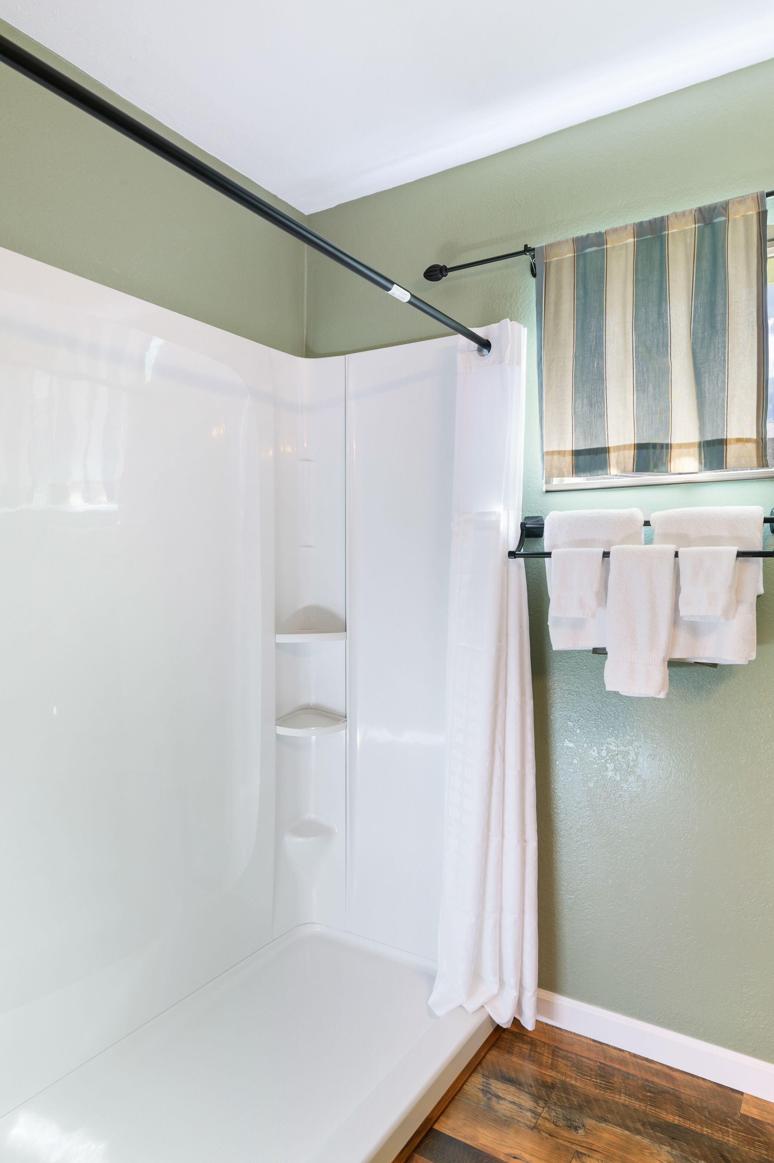 A bathroom with a shower, a towel rack with towels, a window with curtains, and a wooden floor.