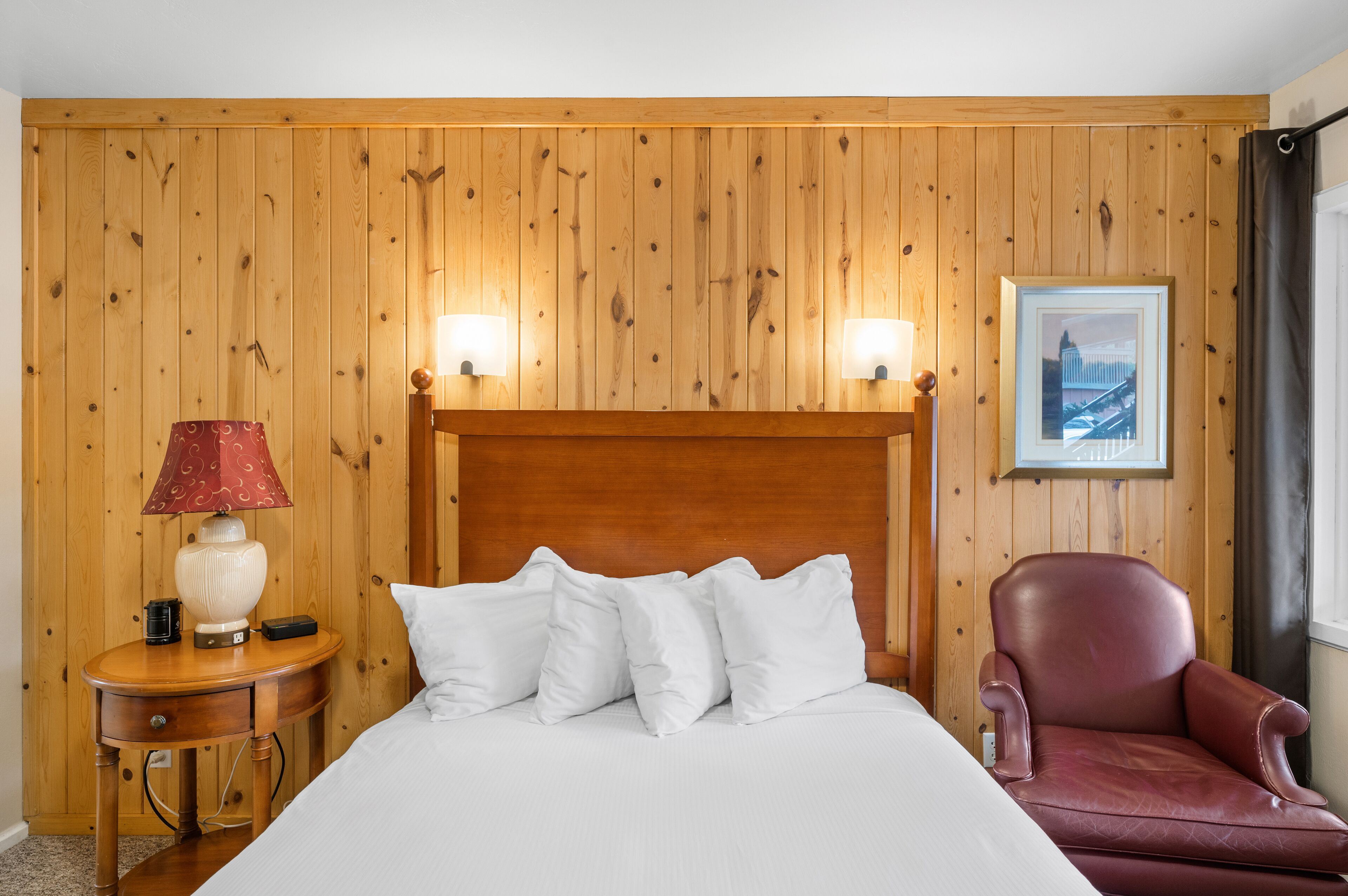 A wooden-paneled room with a bed, a red chair, a nightstand with a lamp, and a framed picture on the wall.