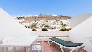 Una terraza con una mesa y sillas blancas, una silla lounge acolchada de color verde, y vistas a un pueblo en la ladera con edificios blancos.