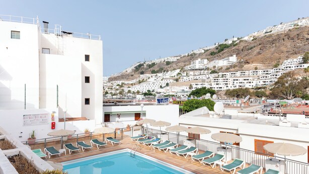 Piscina en la azotea con sillones de descanso y sombrillas, con vistas a una colina de edificios blancos.