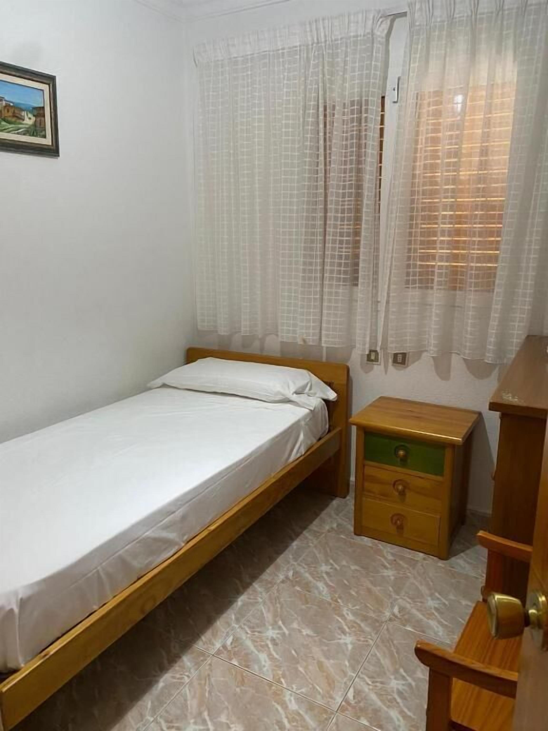 Un lit simple avec un cadre en bois, une petite commode en bois avec des tiroirs et une fenêtre avec des rideaux blancs.