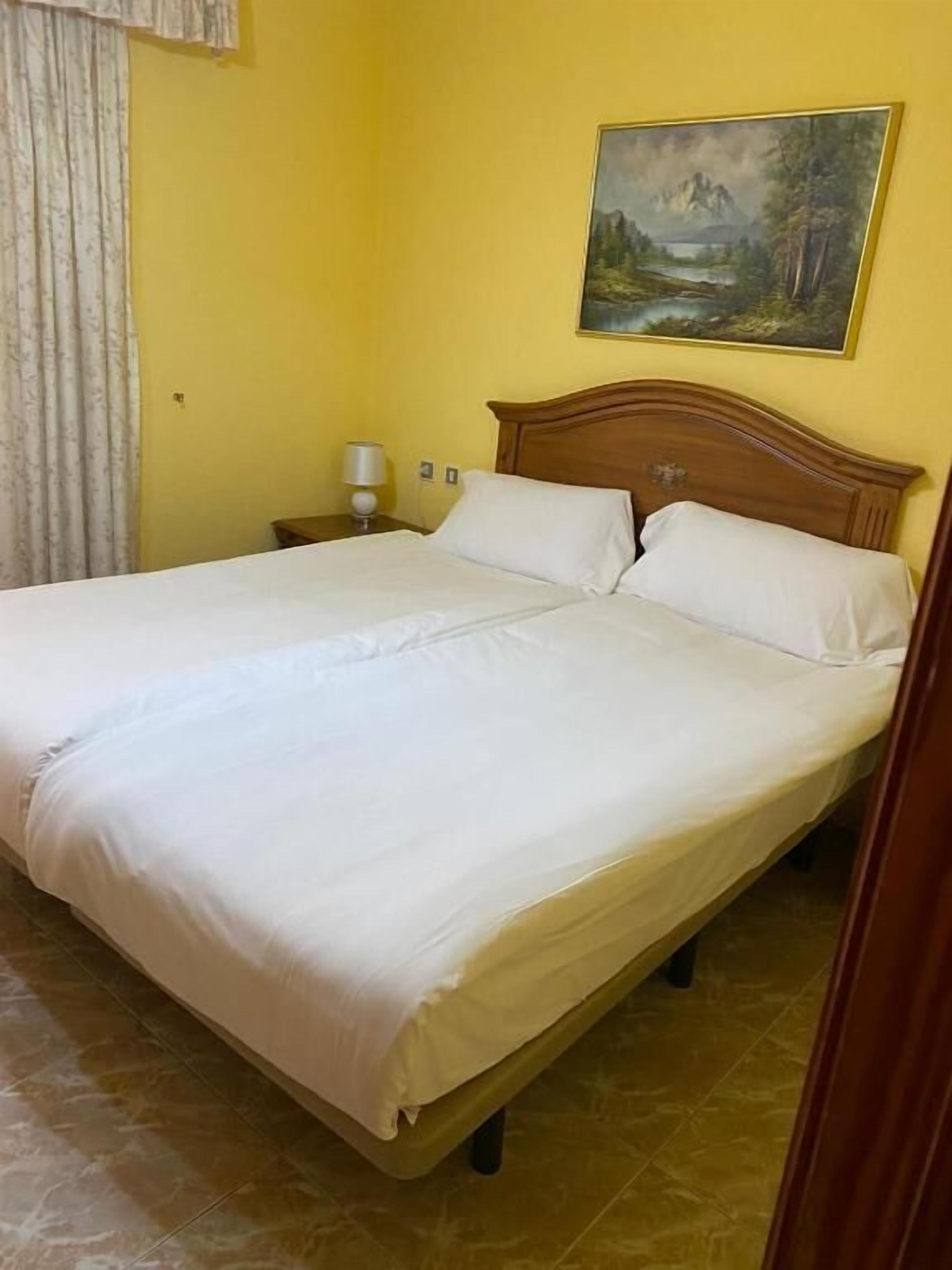 Cama doble con ropa blanca en una habitación con paredes amarillas y un cuadro sobre ella.
