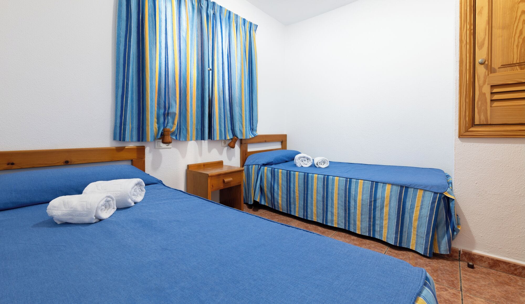 Una habitación con dos camas, cada una con ropa de cama azul y cortinas a rayas. Hay una mesita de noche de madera entre las camas y una puerta de madera a la derecha.