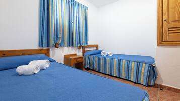Una habitación con dos camas, cada una con ropa de cama azul y cortinas a rayas. Hay una mesita de noche de madera entre las camas y una puerta de madera a la derecha.
