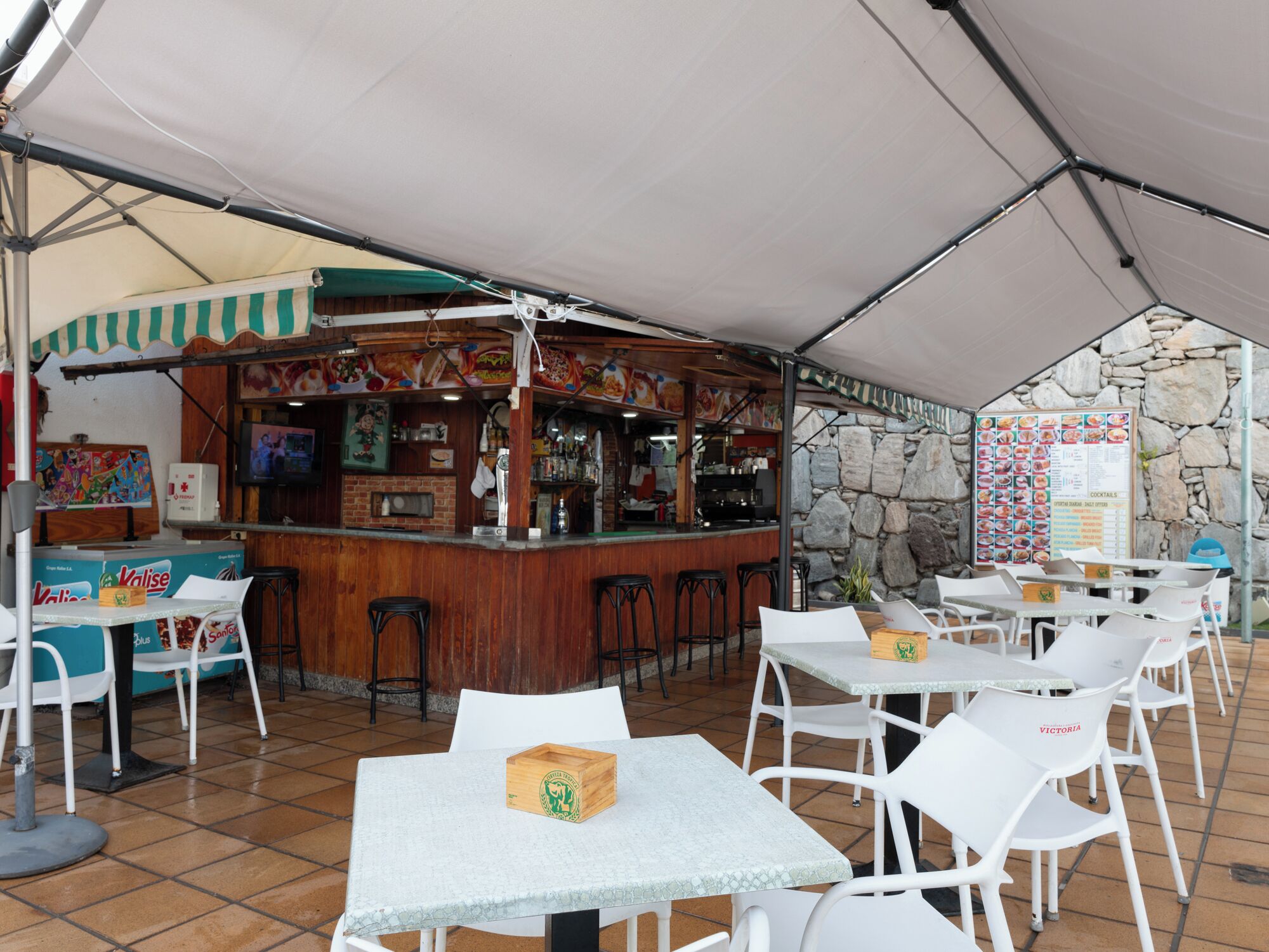 An outdoor café with a bar area, white tables and chairs, and a menu board.