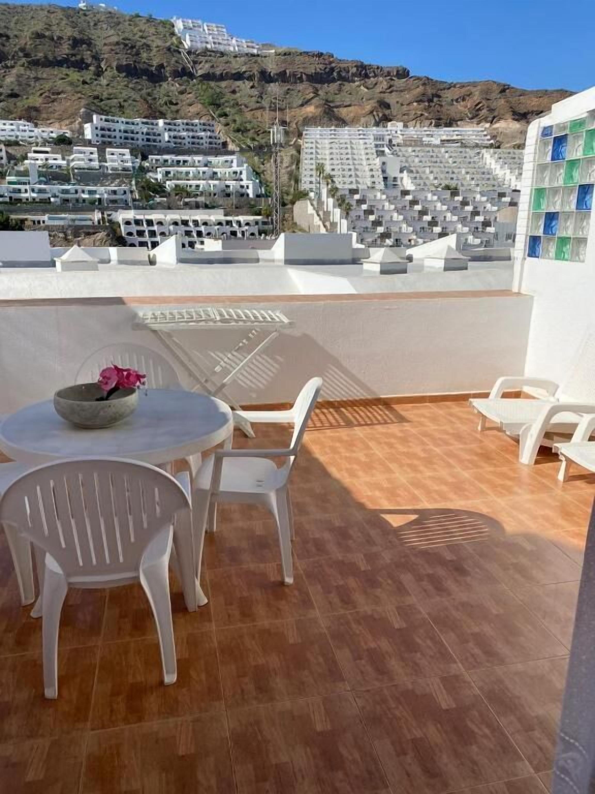 Une terrasse avec une table ronde et des chaises, un saladier rempli de fruits et une vue sur une colline parsemée de bâtiments.