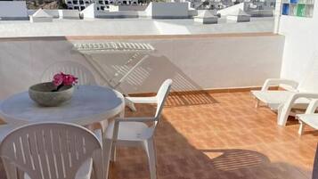 Une terrasse avec une table ronde et des chaises, un saladier rempli de fruits et une vue sur une colline parsemée de bâtiments.
