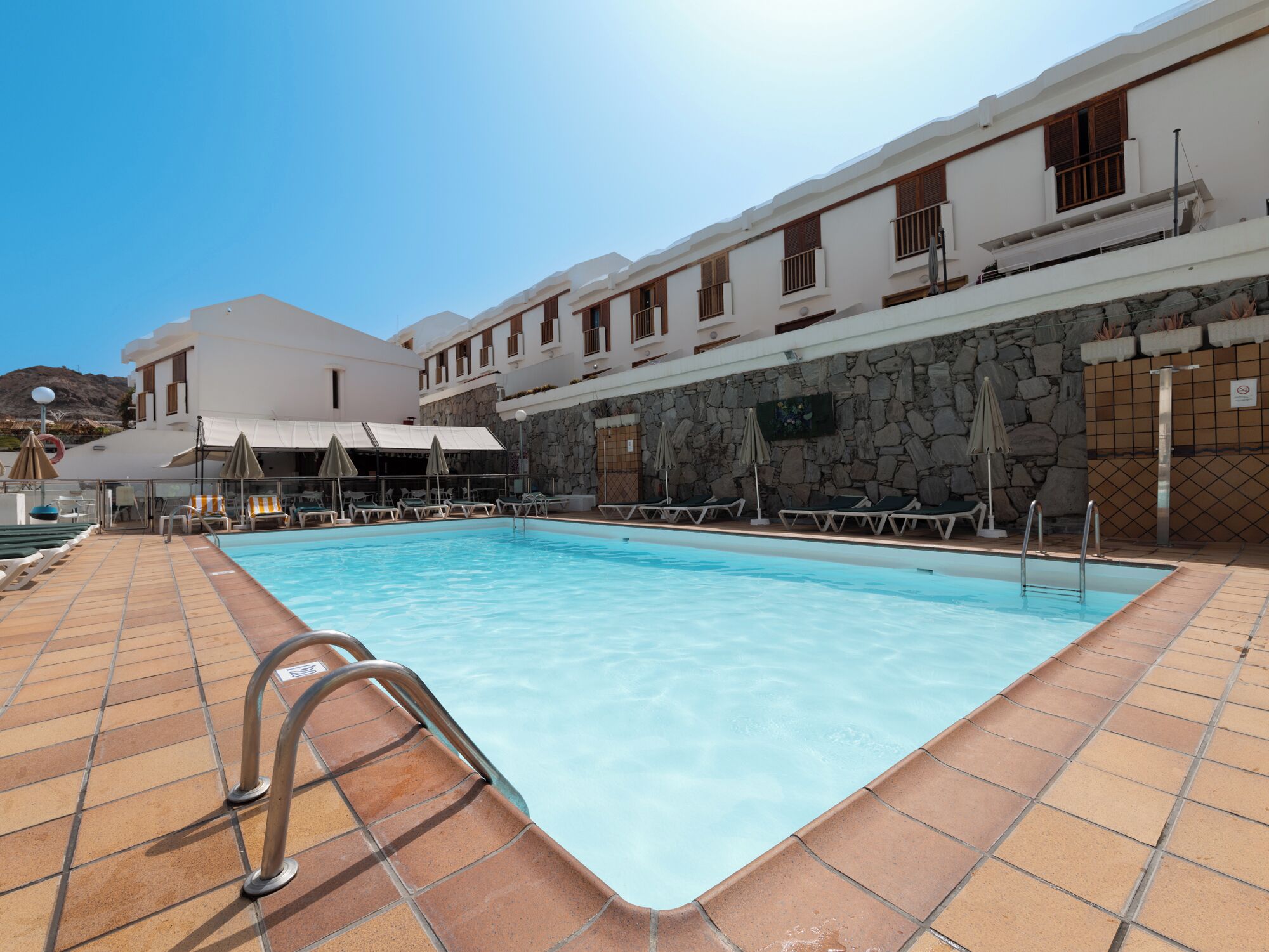 Una piscina con tumbonas y sombrillas, rodeada por un edificio de paredes de piedra y varios balcones.