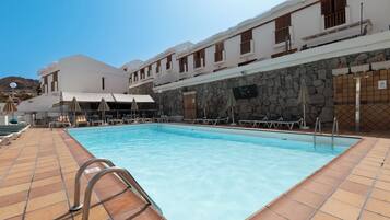 Una piscina con tumbonas y sombrillas, rodeada por un edificio de paredes de piedra y varios balcones.