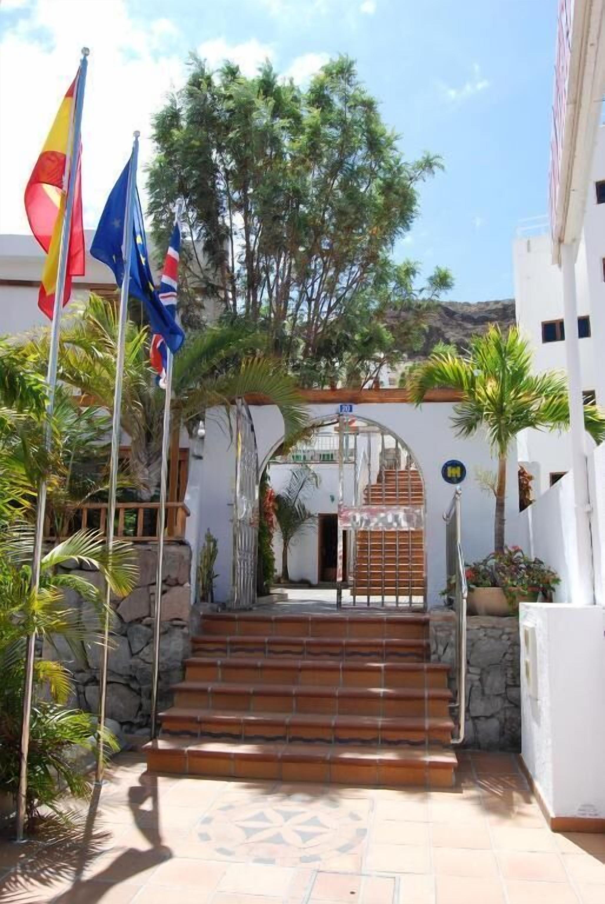 Una entrada de edificio con escalones que conducen a un portón, rodeada de palmeras y banderas.