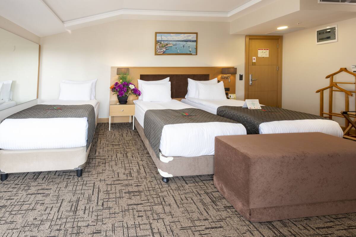 Ein Hotelzimmer mit zwei Betten, einem braunen Hocker, einem Nachttisch mit Lampe und einem Gemälde eines Hafens an der Wand.