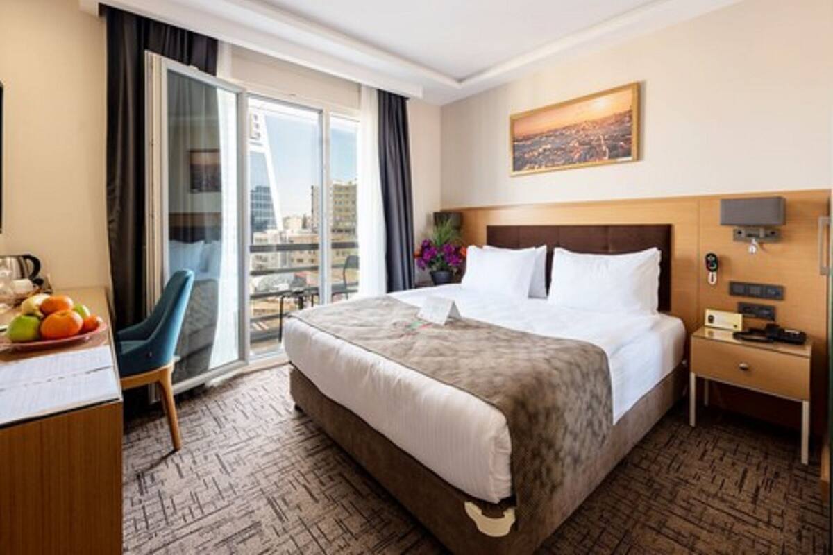 Ein Hotelzimmer mit einem großen Bett, einem Schreibtisch mit Früchten, einem Sessel, einem Nachttisch und Blick auf eine Stadtlandschaft durch eine Glastür.