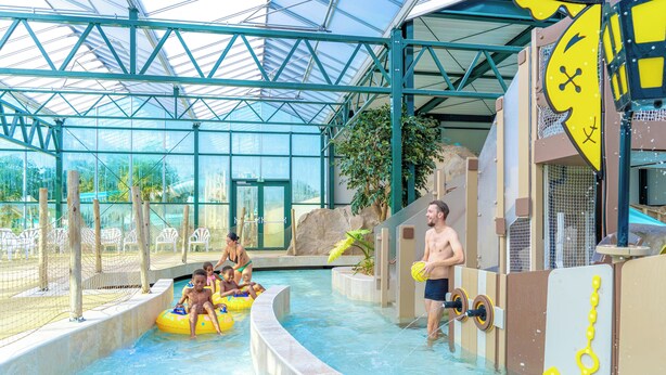 Parque acuático interior con techo de cristal, piscina y estructuras de juego.