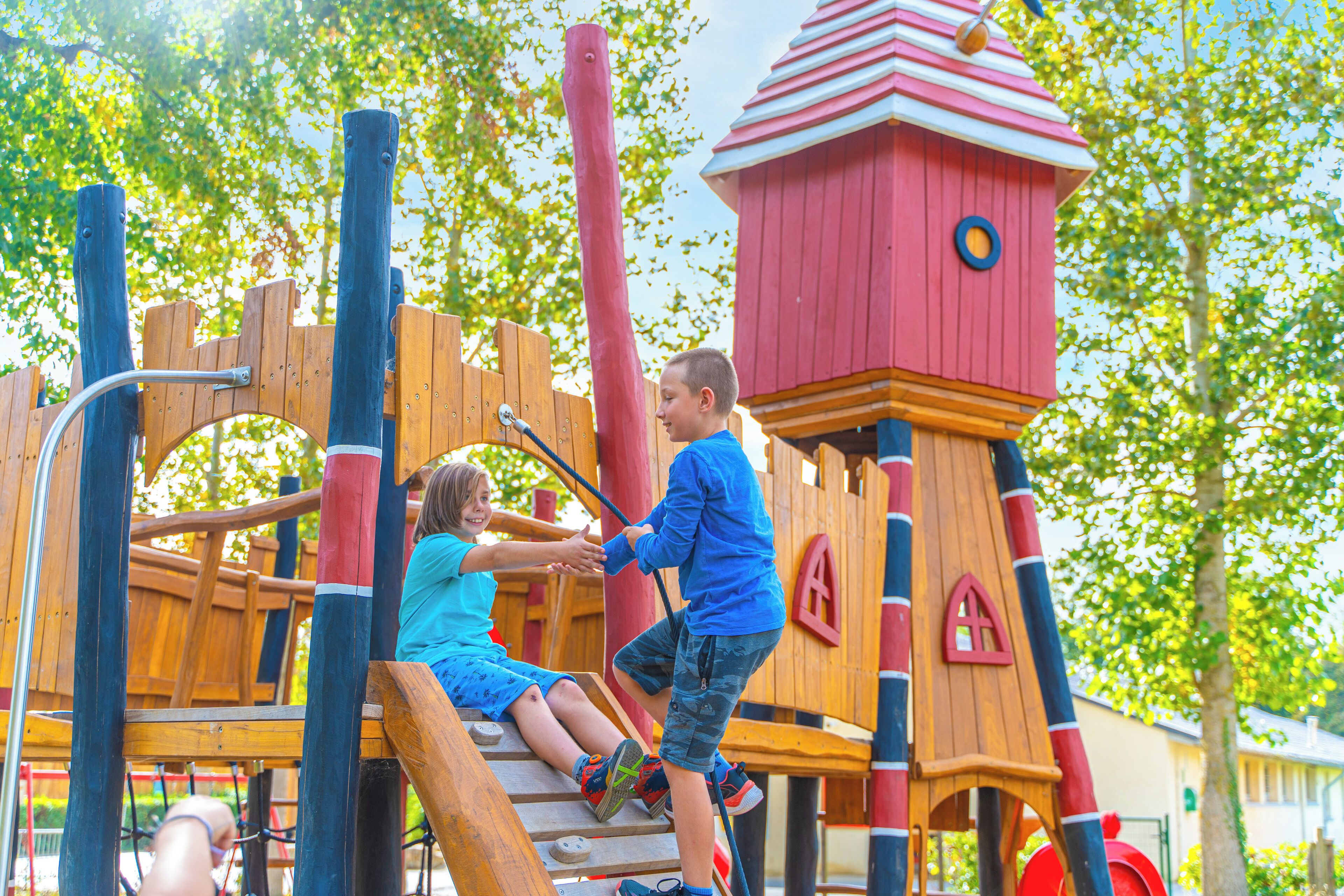 Children playing on a wooden play structure with a red tower and a slide.