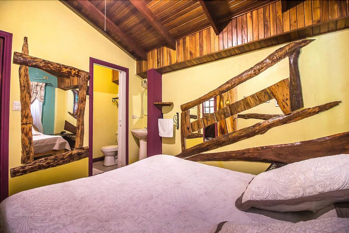 Une chambre à coucher avec une tête de lit en bois, un lit recouvert d’une couvre-lit à motifs, un miroir et une salle de bain équipée de toilettes et d’un lavabo.