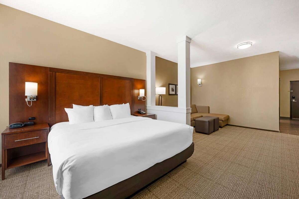 A hotel room with a large bed, a wooden headboard, a nightstand with a lamp, a desk with a chair, and a sofa.