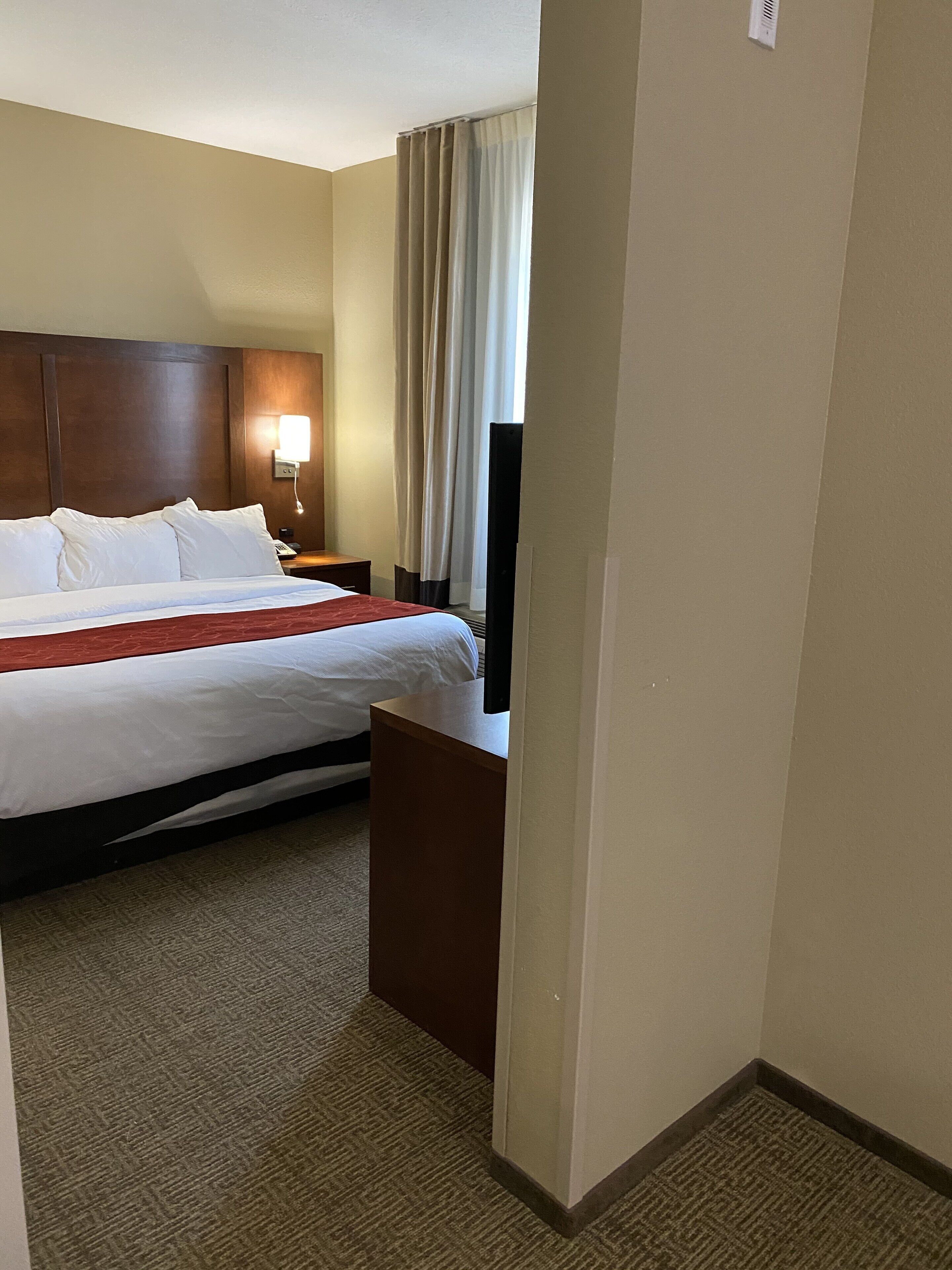 Habitación de hotel con una cama grande, una mesita de noche con lámpara, un televisor de pantalla plana y una ventana con cortinas.