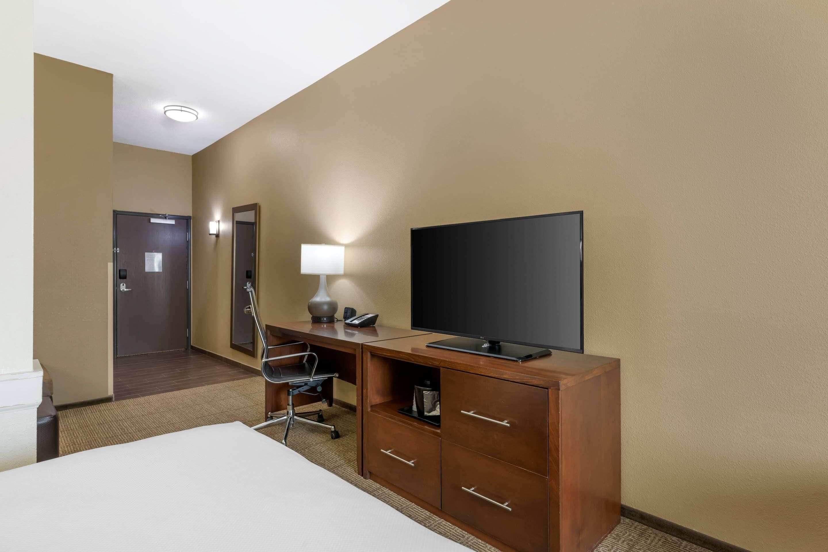 A hotel room with a bed, a desk with a computer, a television, a mirror, and a door.