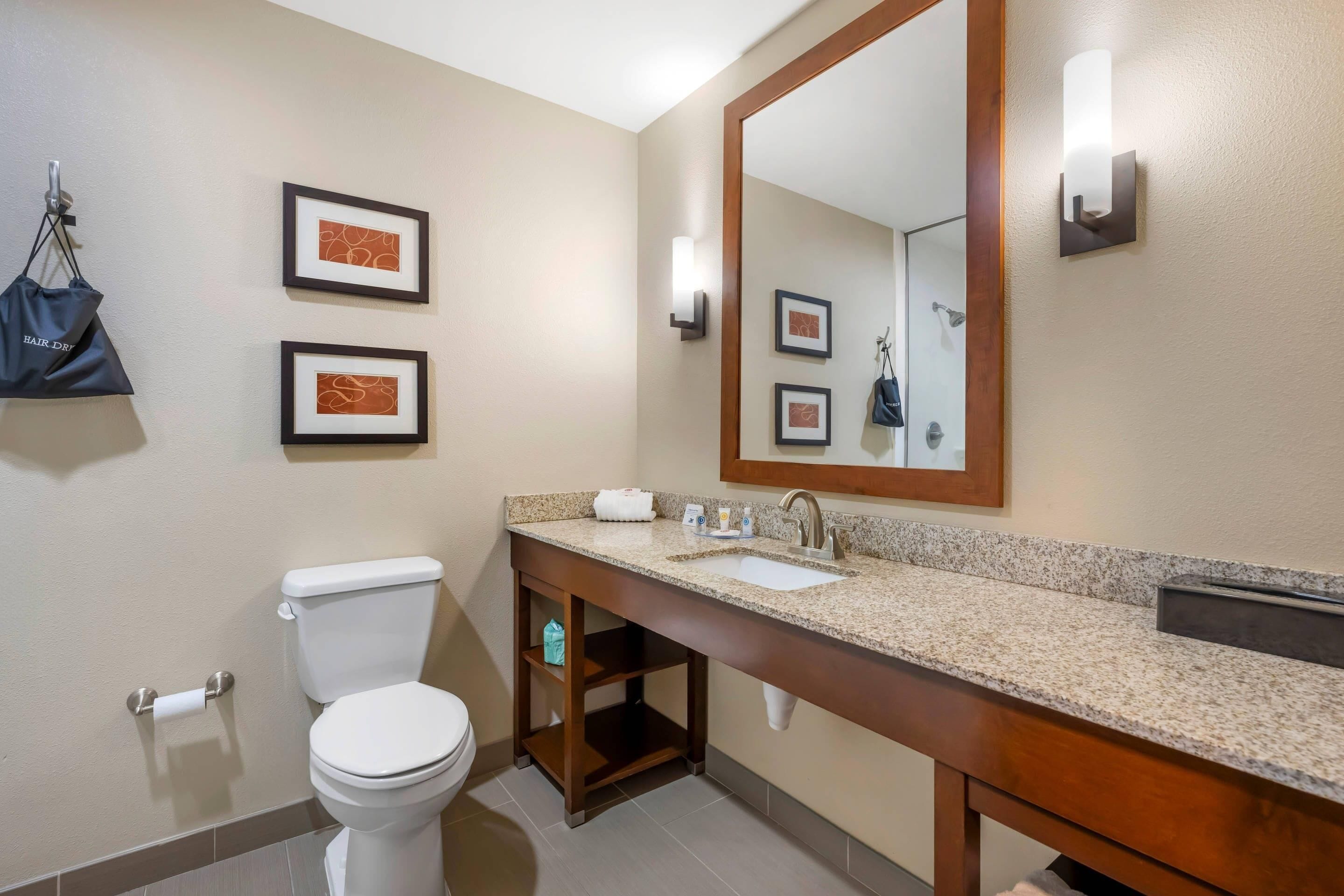 A bathroom with a toilet, sink, and mirror. There are framed pictures on the wall and a bag hanging on the wall.