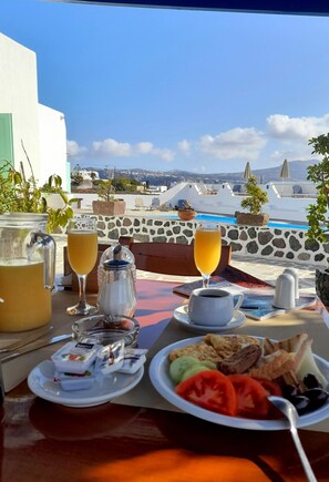 Ein Esstisch mit Blick auf einen Pool und Gebäude, eine Karaffe Orangensaft, eine Kaffeetasse und ein Teller mit Toast, Eiern und Tomaten.