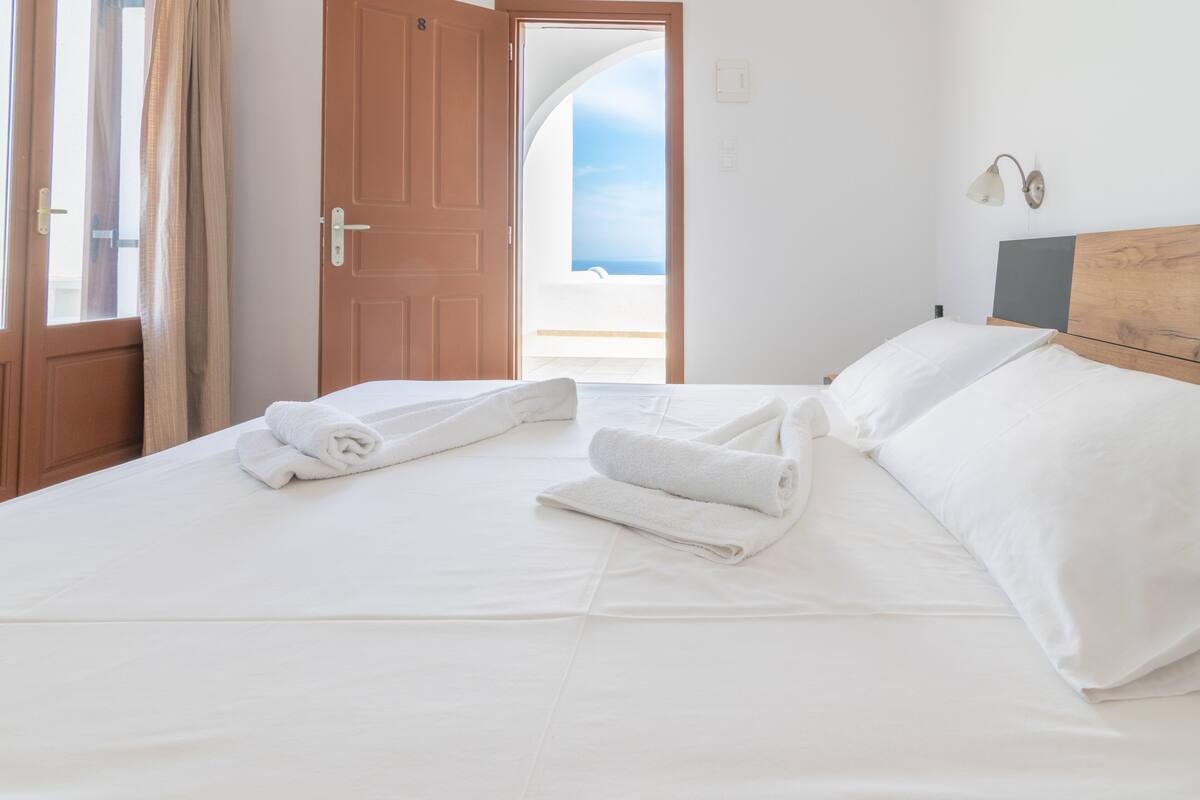 A hotel room with a bed, white linens, a wooden headboard, and a view of the sea through an open door.