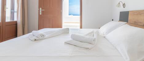 A hotel room with a bed, white linens, a wooden headboard, and a view of the sea through an open door.