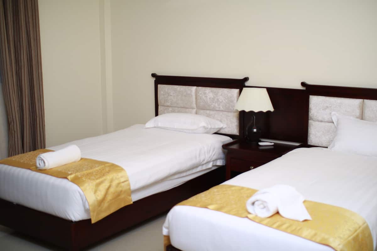 A hotel room with two single beds, each with a white bedspread and a gold accent. There is a wooden nightstand with a lamp between the beds.