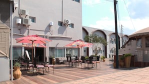 An outdoor patio with tables and chairs, red umbrellas, and potted plants. - The Matola Hotel (Matola)