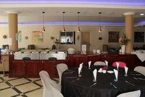 A dining area with a buffet setup, including a stove and various food items. There are tables set with white tablecloths, napkins, and cutlery. The room has wooden cabinets and a checkered floor.