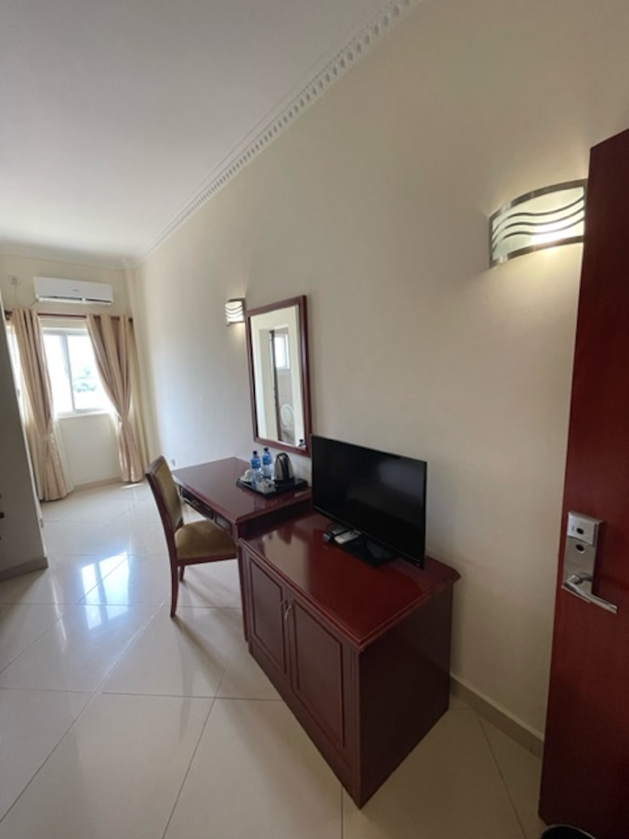 Une chambre d’hôtel avec un bureau en bois, un téléviseur à écran plat, une chaise, un miroir et une porte dotée d’une poignée.