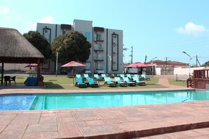 A swimming pool with lounge chairs and umbrellas, a thatched roof structure, and a multi-story building in the background.