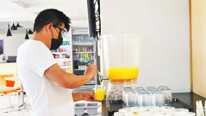 A person pouring orange juice into a glass at a juice bar with a refrigerator and a tray of food items in the background. - Urbainn Hotel (Veracruz)