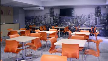 A classroom with orange chairs and tables, a cityscape mural on the wall, and a flat-screen TV mounted above a table.