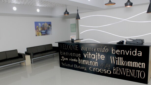 A modern reception area with a black reception desk displaying multilingual welcome messages. There are black sofas and a glass table with a magazine. - Urbainn Hotel (Veracruz)